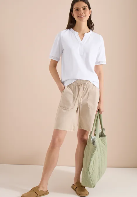 Vrouw in wit T-shirt, beige bermudashort, lichtbruine sandalen en een groene tas.