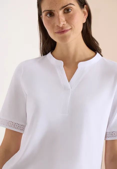 Vrouw in een witte blouse met korte mouwen en subtiele kanten zoom, glimlachend voor een lichte achtergrond.
