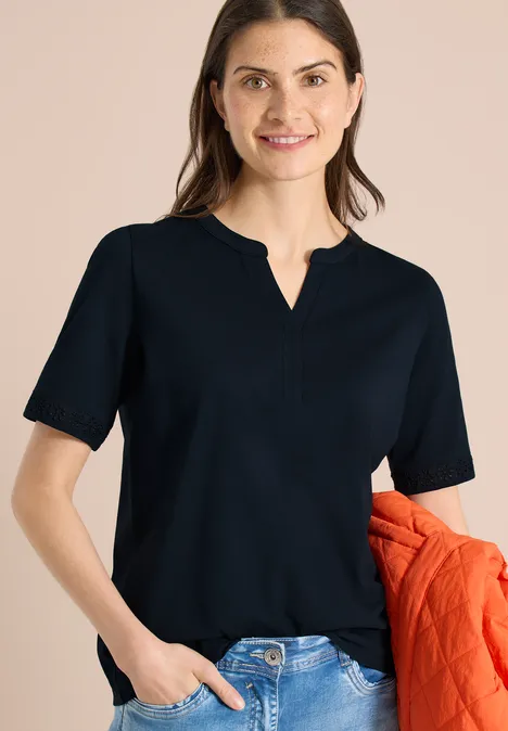 Vrouw in zwart T-shirt met V-hals, spijkerbroek en een oranje jas onder haar arm.