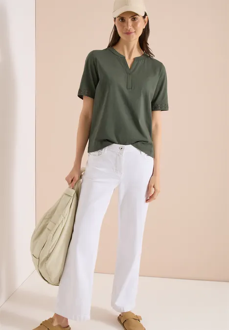 Vrouw in donkergroen V-shirt en witte jeans, met een beige tas, tegen een lichte achtergrond.