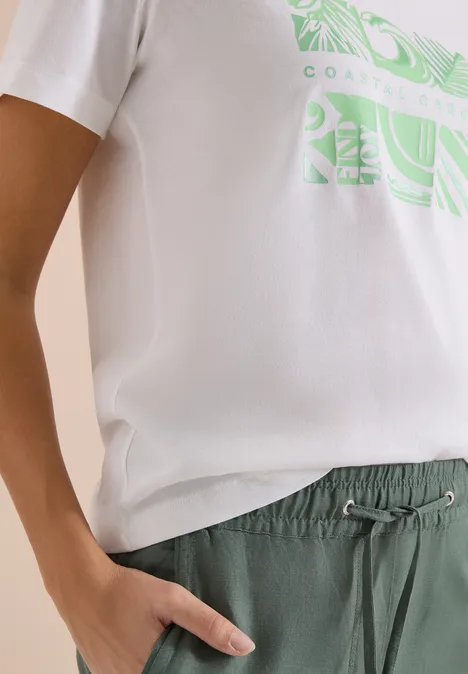 Eine Person trägt ein weißes T-Shirt mit grünem Print und grüne Hose mit Kordel, Hand in Tasche.