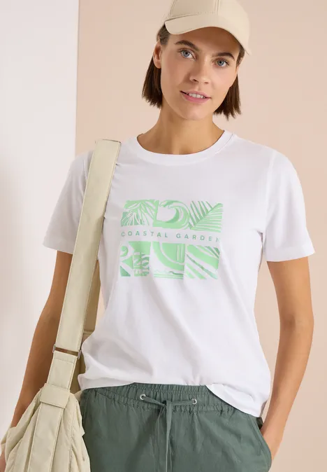 Person trägt weißes T-Shirt mit grünem Print, grüne Hose, Cap und helle Umhängetasche.