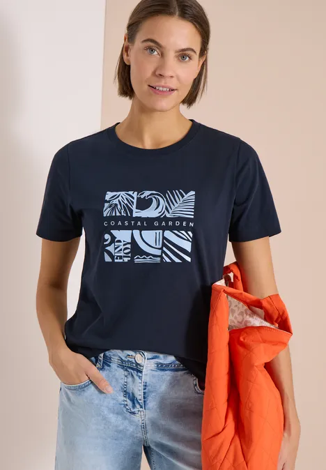 Frau trägt marineblaues T-Shirt mit grafischem Print und hält eine orange Jacke in der Hand.