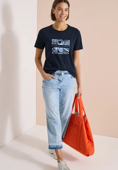 Frau trägt dunkelblaues T-Shirt mit Grafik, locker sitzende helle Jeans und eine orangefarbene Tasche.