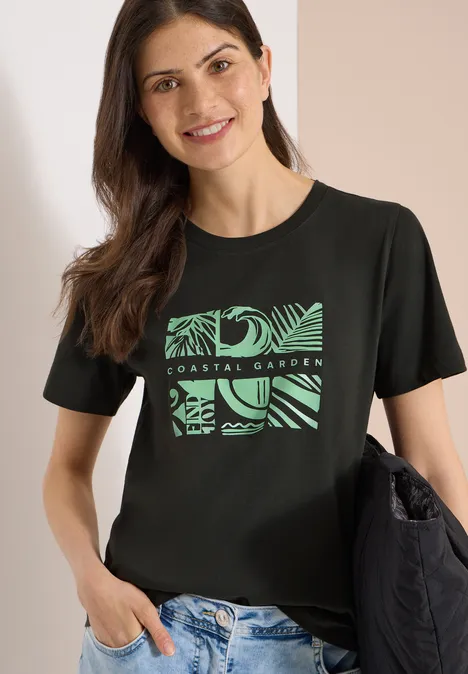 Vrouw met zwart T-shirt met groen plantenprint, lichte spijkerbroek en zwarte schoudertas.