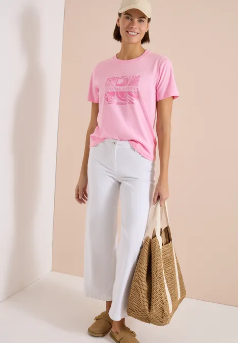 Person trägt rosa T-Shirt mit Print, weiße weite Hose, braune Loafers und hält eine geflochtene Tasche.