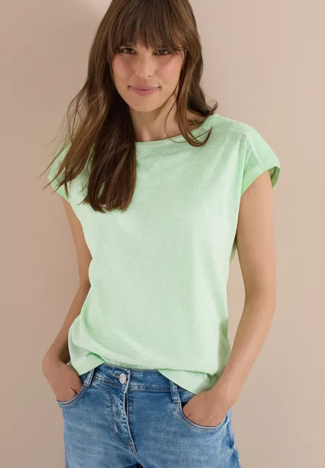 Frau trägt hellgrünes T-Shirt und blaue Jeans, steht mit Händen in den Taschen.