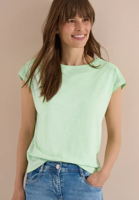 Frau in mintgrünem T-Shirt und blauen Jeans vor beigem Hintergrund, eine Hand in der Hosentasche.