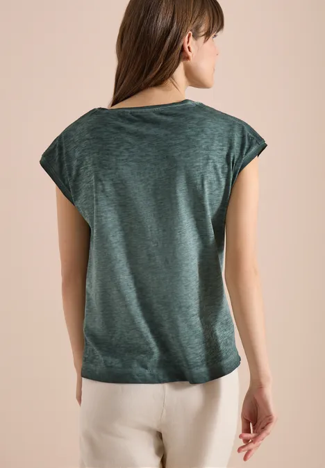 Eine Frau von hinten trägt ein grün meliertes T-Shirt mit überschnittenen Schultern und beige Hose.