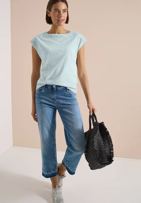T-Shirt aus Jersey im Garment Dye Look crystal mint Frau mit hellblauem T-Shirt, blauen Jeans und schwarzer geflochtener Tasche vor neutralem Hintergrund.