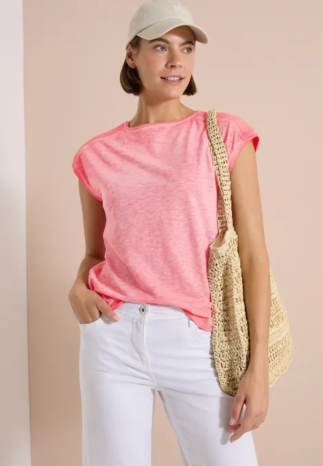 T-Shirt aus Jersey im Garment Dye Look coral Frau im pinken T-Shirt und weißen Jeans mit Strohtasche und Baseballcap vor hellem Hintergrund.