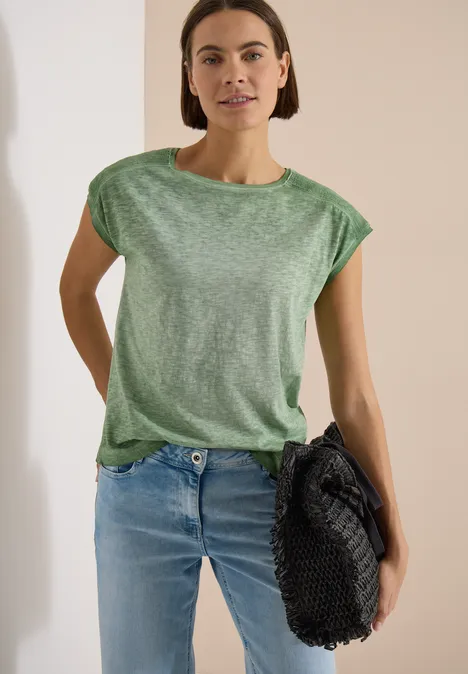 Een persoon staat met een groen shirt, een spijkerbroek en houdt een zwarte tas vast.