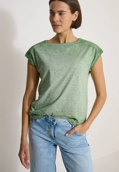 Een vrouw in een los groen T-shirt en blauwe jeans, met één hand in de zak.