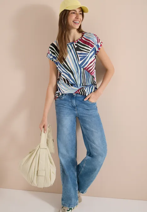 Person trägt bunte gestreifte Bluse, weite Jeans, gelbe Cap und große beige Schultertasche.