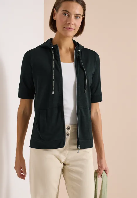 Frau trägt schwarzen Kurzarm-Hoodie mit Reißverschluss, weiße Bluse, helle Hose, hält grünen Pullover.