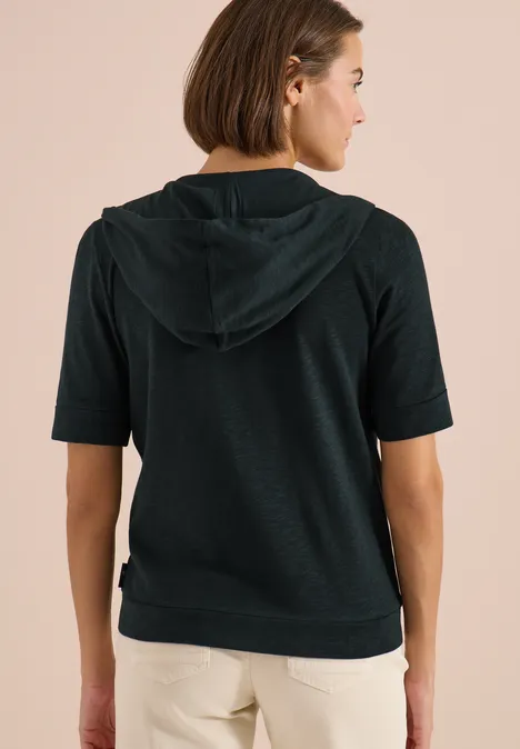 Frau von hinten in dunkelgrauem kurzärmeligem Kapuzenpullover und hellen Hosen.