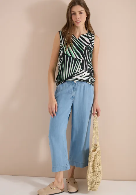 Frau trägt gestreiftes Top, weite blaue Culotte und Basttasche vor neutralem Hintergrund.