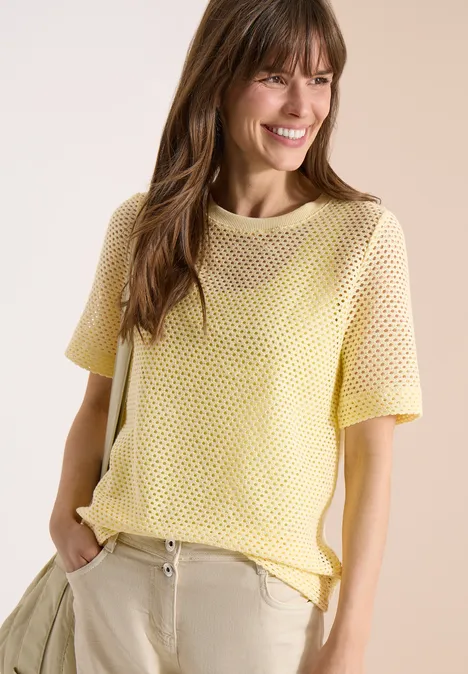 Frau mit gelbem Netzt-T-Shirt und beigen Hosen, lässig mit den Händen in den Taschen stehend.