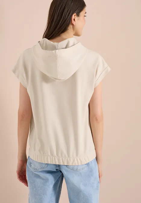 Frau von hinten in beiger Kapuzenbluse mit kurzen Ärmeln und lockerer Passform vor hellem Hintergrund