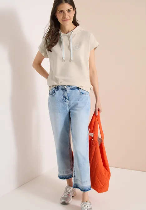 Frau im beigen Kapuzen-Shirt und weiten, hellblauen Jeans, die eine orangefarbene Tasche trägt.