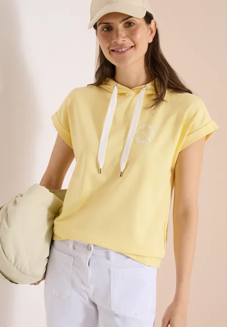 Frau in gelbem Kurzarm-Hoodie mit weißer Kordel, weiße Jeans, hält eine passende Tasche.