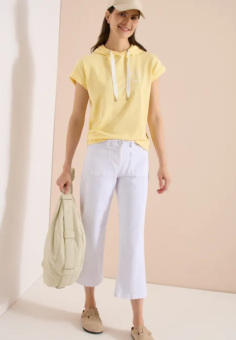 Junge Frau in gelbem Kapuzen-Shirt, weißer Schlaghose, beiger Kappe und Slippern hält gesteppte Tasche.