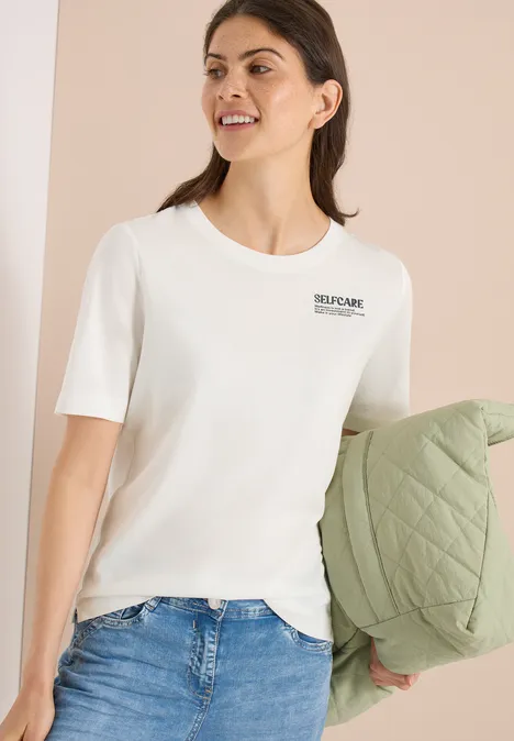 Vrouw draagt een wit T-shirt met korte mouwen en houdt een groen doorgestikt jack vast.