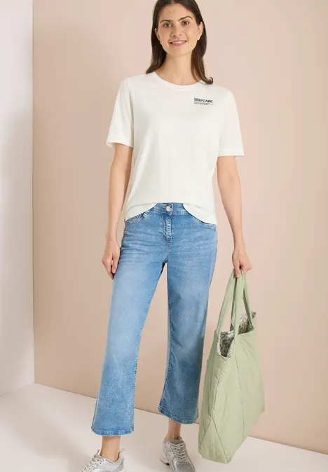 Vrouw in wit T-shirt, blauwe jeans en grote groene tas tegen een neutrale achtergrond.