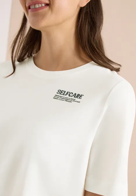 Een vrouw draagt een wit shirt met een kleine 'SELF-CARE'-opdruk.