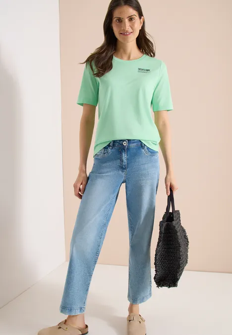 Persoon draagt lichtgroen T-shirt, rechte lichtblauwe jeans en houdt een zwarte tas vast.