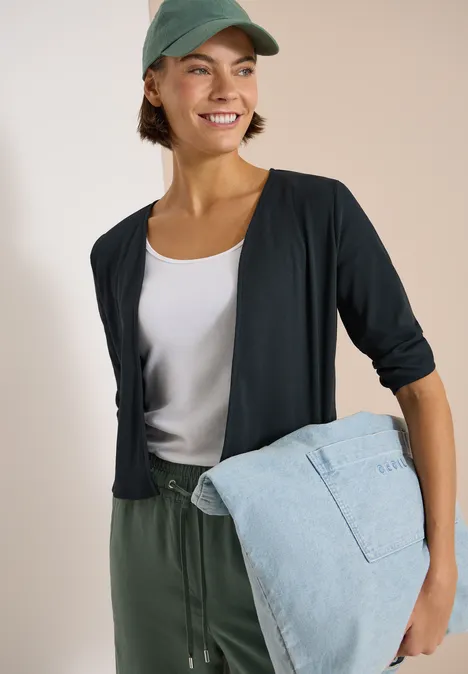 Person trägt weißes Top, schwarzen Kurzcardigan, grüne Hose und hält eine hellblaue Jeansjacke.