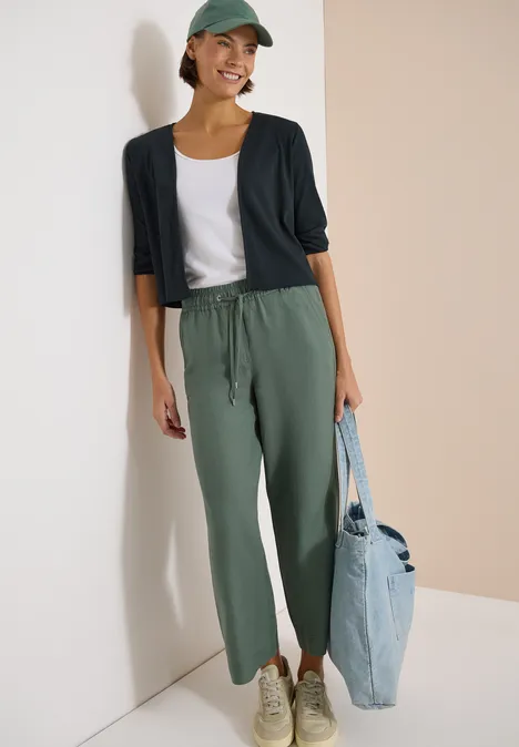 Eine Person steht vor einer Wand, trägt grüne Jogginghose, weißes T-Shirt, dunkelblauen Cardigan, Cap und hellblaue Tasche.