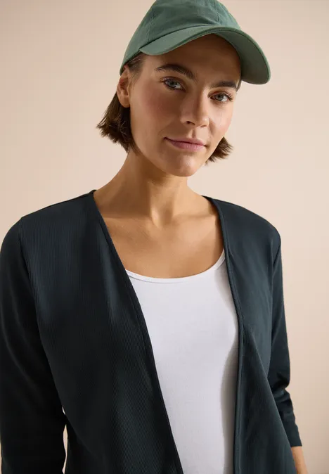 Oberkörper einer Person in schwarzer Jacke, weißem Top und grüner Cap, Gesicht unkenntlich.