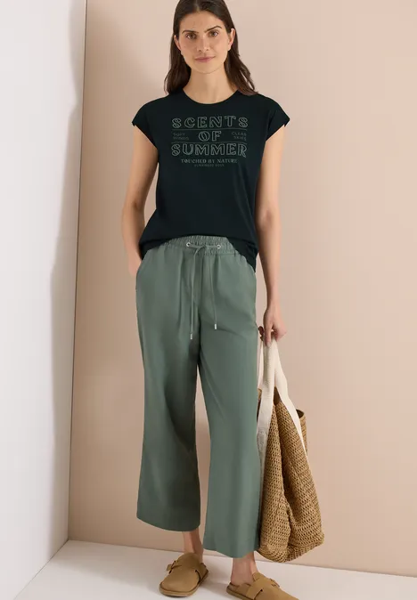 Model draagt zwart t-shirt met print, wijde groene broek en heeft een gevlochten strooien tas.
