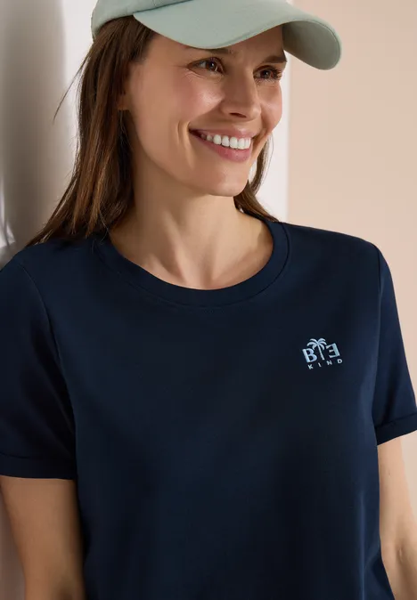 Frau mit Baseballcap und dunkelblauem T-Shirt mit kleinem Logo vor heller Wand.