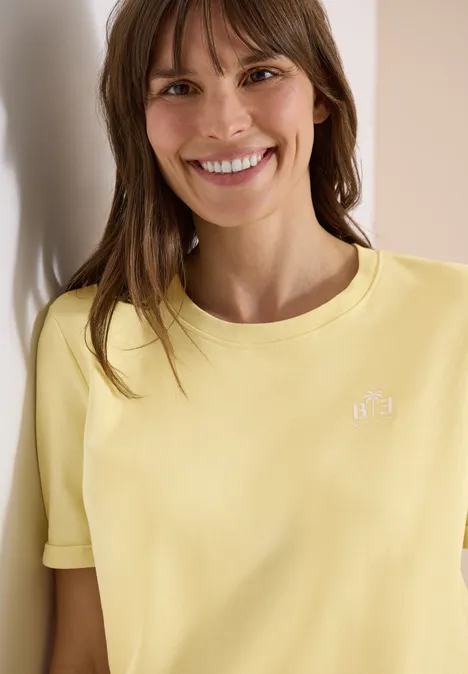 Eine Frau steht an einer Wand und trägt ein hellgelbes T-Shirt mit kleinem weißen Logo.