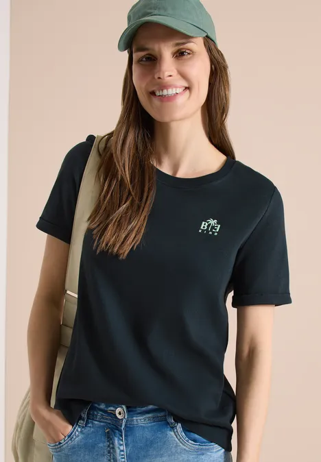 Femme en t-shirt noir à petit logo, jean, casquette et sac épaule devant fond clair