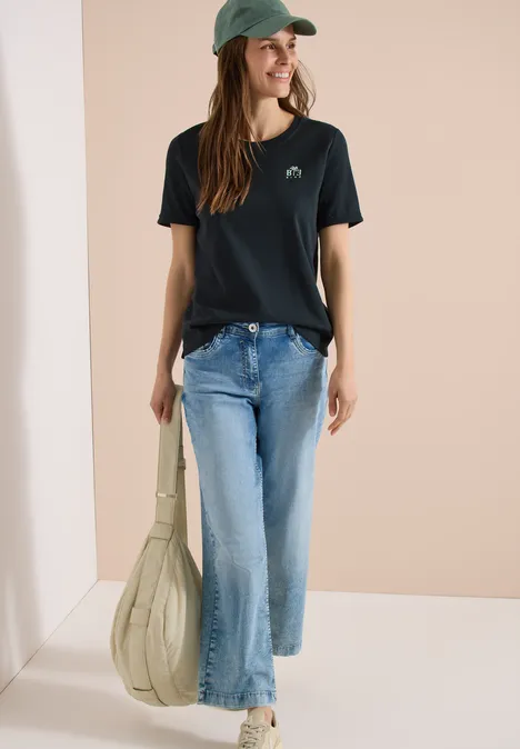 Femme en t-shirt noir avec motif baleine, jean bleu, casquette verte, baskets blanches et grand sac vert clair.