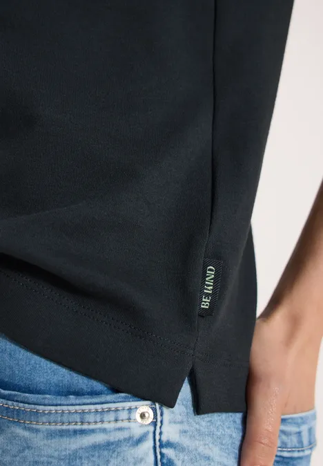 Gros plan d’un t-shirt noir avec fente latérale et petite étiquette logo, porté avec un jean bleu.