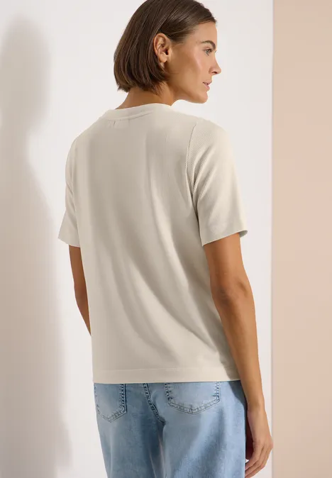 Rückenansicht einer Person mit beigem T-Shirt und hellen Jeans vor neutralem Hintergrund.