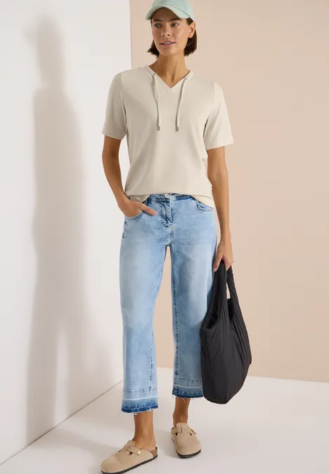 Person trägt kurzes Kapuzen-T-Shirt, weite, blaue Cropped-Jeans, beige Slipper und hält eine schwarze Tasche.