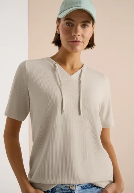 Person trägt hellbeiges, kurzärmeliges Kapuzen-Shirt vor neutralem Hintergrund.