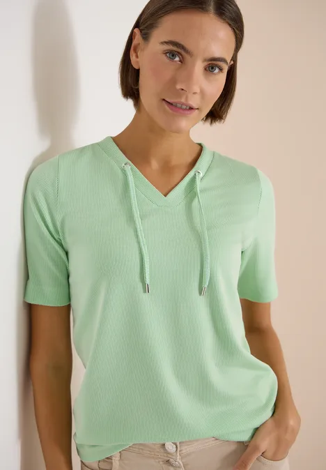 Frau trägt mintgrünes Kurzarm-Shirt mit Kapuze und V-Ausschnitt, steht entspannt mit einer Hand in der Tasche.