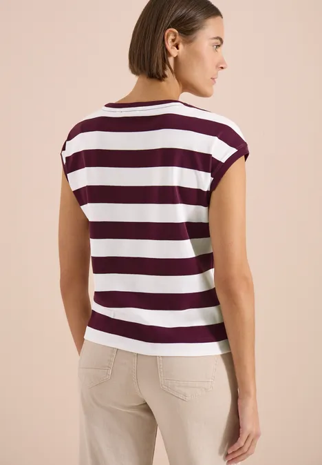 Een vrouw staat met haar rug naar de camera en draagt een wit-bordeaux gestreept shirt met beige broek.