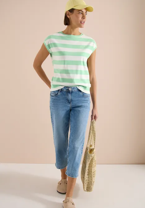 Vrouw in groen-wit gestreept T-shirt, lichtblauwe cropped jeans, sandalen en rieten tas.