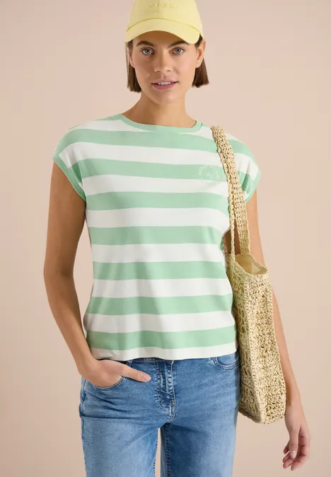 Dame draagt een groen-wit gestreept T-shirt, spijkerbroek en een gevlochten strooien tas over de schouder.