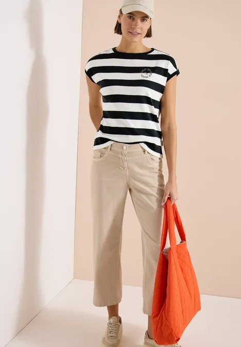 Vrouw met zwart-wit gestreept shirt, beige broek, sandalen en een oranje tas voor neutrale achtergrond.