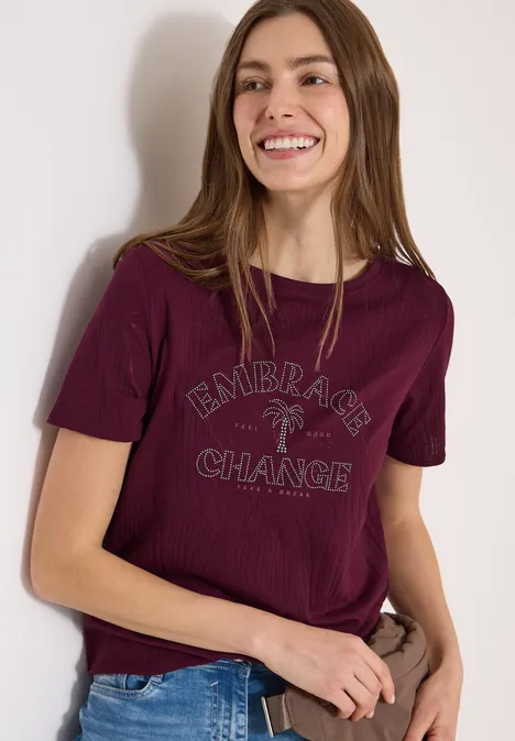 Frau trägt bordeauxrotes T-Shirt mit „Embrace the Change“-Aufdruck und Palmenmotiv, hält geflochtene Tasche.