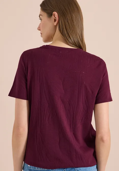 Frau von hinten in bordeauxrotem T-Shirt vor neutralem hellen Hintergrund.