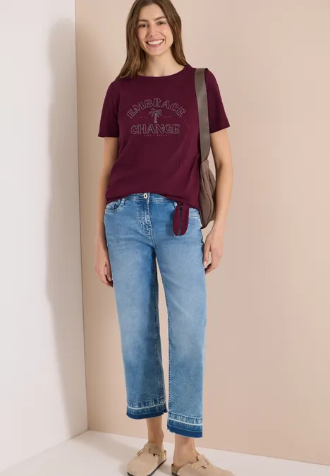 Frau trägt Bordeaux-T-Shirt, hellblaue Jeans und Beige-Slipper, steht vor beigem Hintergrund mit Schultertasche.